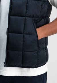 Marineblaue Steppweste mit einem gesteppten Design, Reißverschluss und seitlichen Taschen. Hergestellt aus glänzendem, atmungsaktivem Material mit einer glatten Textur.