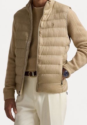 Uomo che indossa un gilet trapuntato beige sopra un maglione marrone chiaro, pantaloni crema, una cintura nera e un orologio scuro, in piedi con una mano in tasca.