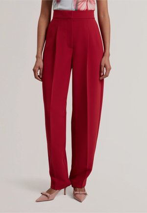 Pantaloni lunghi rossi a vita alta a gamba larga, indossati con tacchi a punta rosa pallido con fiocchi, mostrati dalla vita ai piedi su uno sfondo semplice.