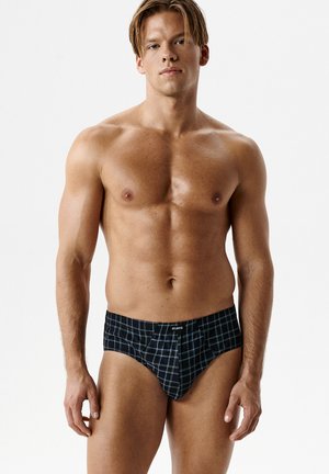Męski mężczyzna o jasnej karnacji i krótkich blond włosach nosi czarne bokserki z białym i niebieskim wzorem w kratę na białym tle.