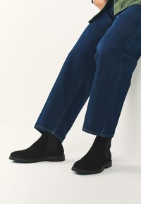 Melni zamšādas potīšu zābaki ar elastīgām sānu panelēm un zemu zoli, kombinēti ar tumši ziliem džinsa biksēm, kas ir saīsinātas pie potītes.