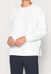 Sudadera blanca con cuello redondo, diseño de logo en relieve en la parte frontal, puños acanalados y aberturas laterales. El material parece suave y texturizado.