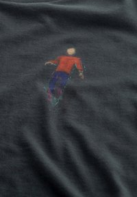 Grijze t-shirt met een afbeelding van een figuur in een rood shirt en blauwe broek. Zachte textuur met een iets relaxte pasvorm.