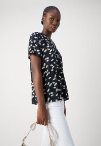 Blusa blu navy a maniche corte con motivo floreale giallo e bianco, dotata di scollatura a V e vestibilità ampia. Abbinata a pantaloni bianchi.