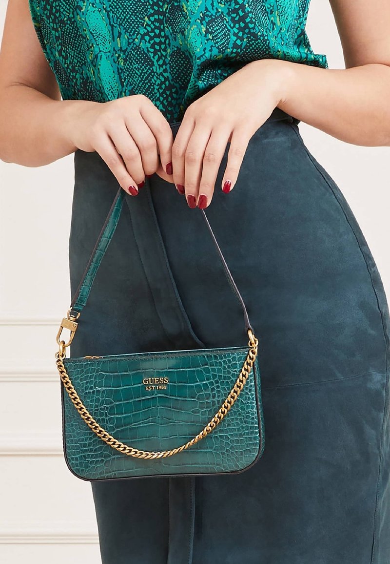 Teal handtas met krokodillenstructuur en een gouden kettingaccent, voorzien van een gegraveerd logo en een afneembare schouderband, naast een suède outfit.