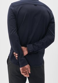 Marinblå långärmad skjorta med mjuk textur, knäppta manschetter och ryggparti med detaljer. Stylad med mörka byxor, vy bakifrån.