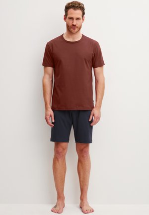 Mann steht barfuß an einer weißen Wand und trägt ein braunes T-Shirt mit kurzen Ärmeln und dunkelgraue kniehohe Shorts.