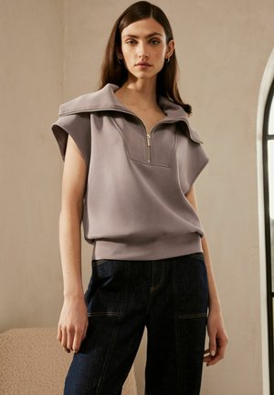 Lyse lilla, kortærmet pullover med høj krave og lynlås, lavet af blødt stof, parret med mørke denimbukser.