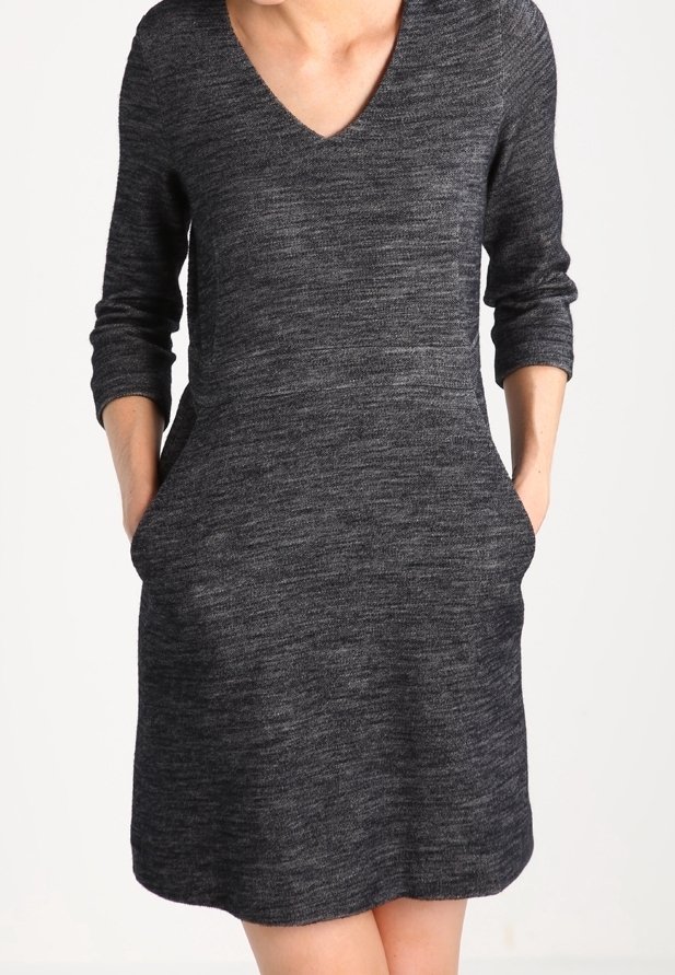 Femme portant une robe en tricot gris foncé à manches longues avec un col en V et les mains dans les poches latérales, debout devant un fond blanc uni.