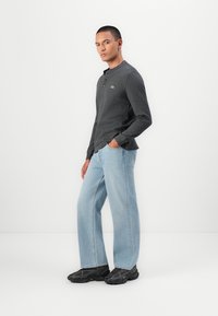 Cămașă henley cu mânecă lungă gri, cu un mic logo verde, asortată cu blugi largi de culoare albastru deschis și adidași negri. Design simplu, croială lejeră.