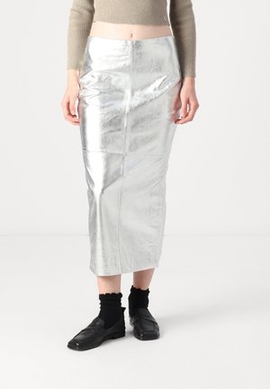 Kvinde iført en metallisk sølv midi-nederdel, beige cropped sweater, sorte sokker og sorte loafers, stående på en hvid gulv og baggrund.