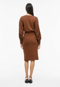 Brązowa dzianinowa sukienka z balonowymi rękawami, dopasowanym krojem i prążkowaną talią. Gładka faktura, długość do kolan, zestawiona z czarnymi sandałami na obcasie.