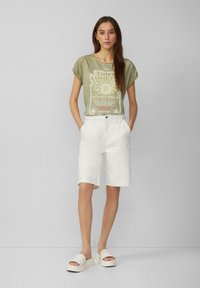 T-shirt olive vert à manches courtes avec imprimé graphique ; bermudas en denim blanc ; sandales compensées beiges. Coupe simple et décontractée.