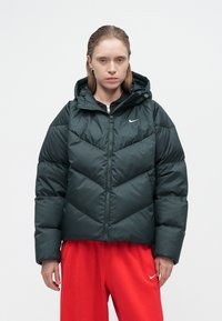 Tamsiai žalia pūkinė striukė su kapisonu, su chevrono raštu, užtrauktuko uždarymu ir subtiliu Nike logotipu. Užsidėta ant juodos bliuskos ir raudonų kelnių.