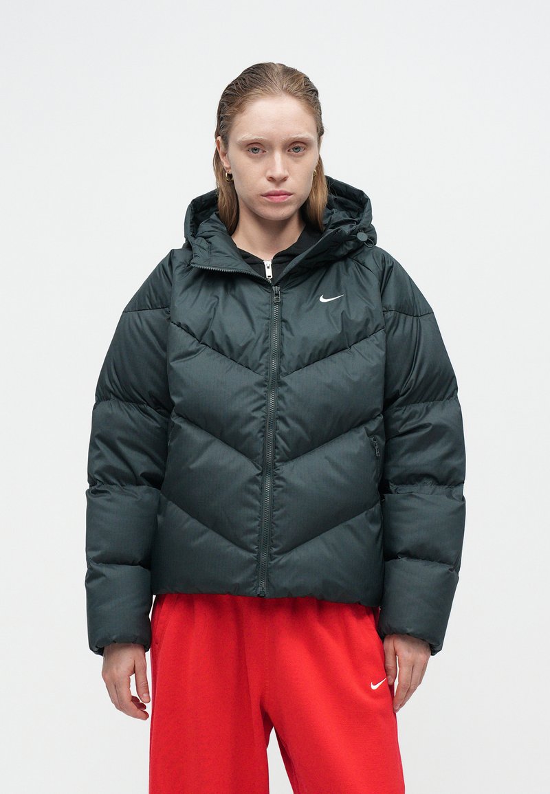 Dark green puffer jacket with a hood, featuring a chevron pattern, zip closure, and subtle Nike logo. Worn over a black top and red pants.