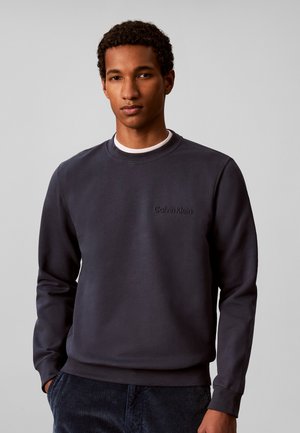 Młody mężczyzna ubrany w czarną bluzę Calvin Klein i ciemne spodnie, stoi z rękami w kieszeniach na jednolitym szarym tle.
