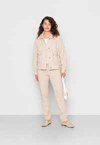 Casaco texturizado bege com dois bolsos frontais, combinado com uma blusa off-white, calças bege e sapatos de cor clara. Uma bolsa branca é segurada na mão.