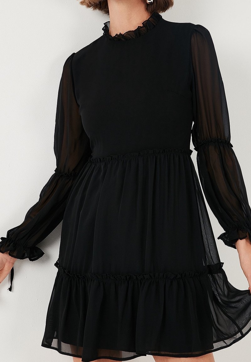 Femme portant une robe noire à manches longues avec des manches transparentes, des poignets à volants, un col montant, une taille froncée et un ourlet à volants.