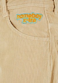 Pantaloni in corduroy beige con righe verticali; presentano una tasca laterale con il logo "homeboy x-tra" ricamato in giallo e blu.