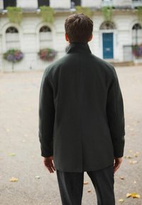 Tmavozelený vlnený kabát s vysokým golierom, priamym strihom a jemnou textúrou. Nosí sa s tmavými nohavicami, na pozadí rozmazaného mestského prostredia.