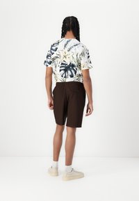 Camisa de manga corta con un patrón floral tropical en verde, amarillo y blanco, combinada con pantalones cortos plisados marrones y zapatillas beige.