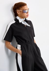 Svart jumpsuit med korte ermer, glidelås foran og sidelommer. Har kontrasterende hvite sidepaneler. Tilbehør inkluderer et hvitt skjerf og solbriller.