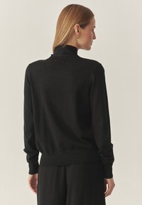 Femeie cu păr drept blond, purtând cercei de aur, un pulover negru cu guler înalt și pantaloni negri, întoarsă lateral, pe un fundal simplu.