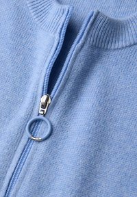 Jasnoniebieski sweter dzianinowy z wysokim kołnierzem i srebrnym zamkiem błyskawicznym. Posiada okrągły, niebieski uchwyt zamka oraz teksturowany detal materiału.