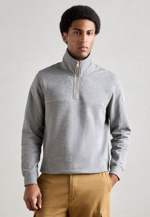 Grauer, Reißverschluss-Sweatshirt mit hohem Kragen. Hergestellt aus weichem Stoff, mit einem nahtlosen Design auf der Brust und gerippten Bündchen. Der Arm hat ein kleines Logo-Detail.