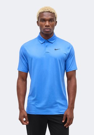 Uomo con capelli biondi e ricci che indossa una polo Nike blu acceso e pantaloni neri, in piedi davanti a uno sfondo chiaro e semplice.