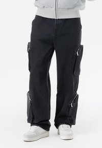 Black denim cargo pants with side zip pockets, relaxed fit, cuffed hems, and contrast stitching. Paired with white sneakers.