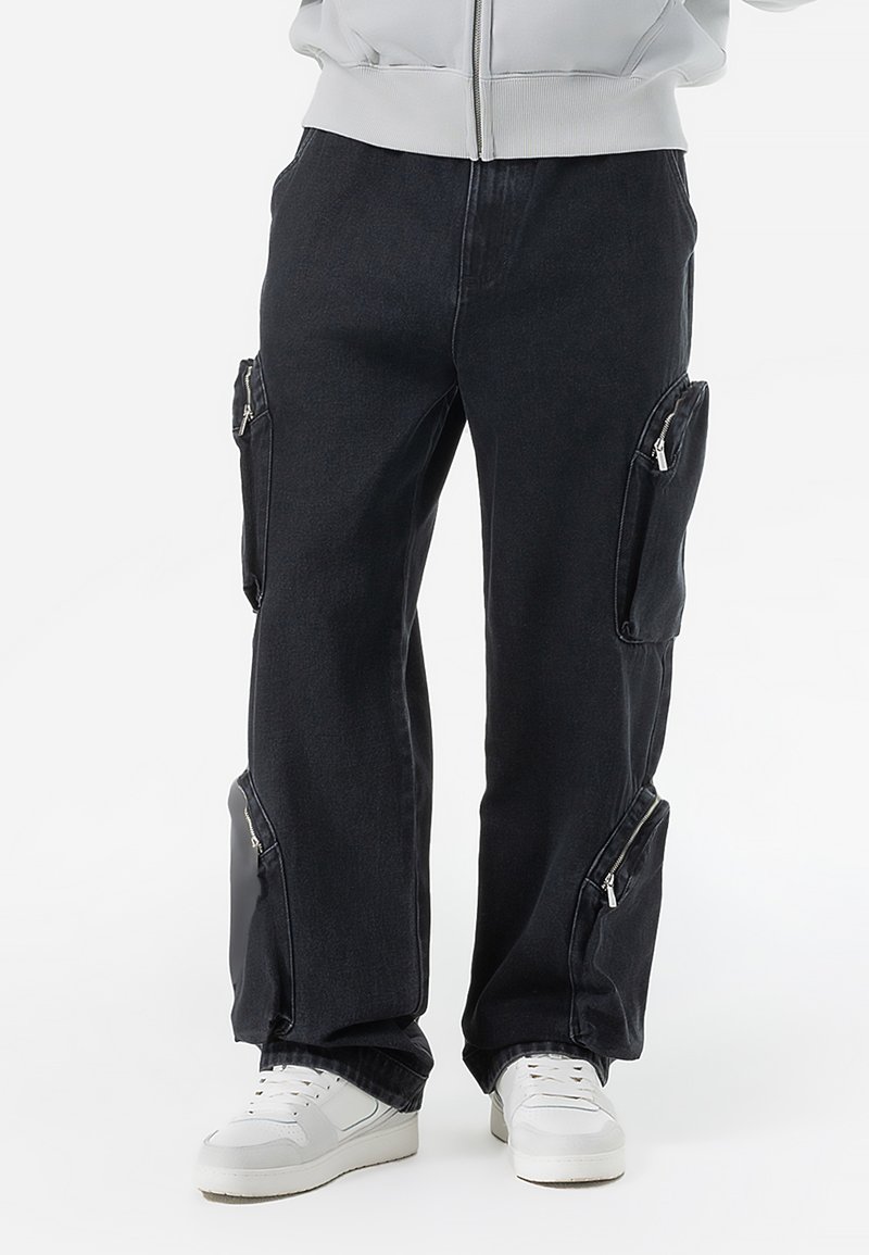 Black denim cargo pants with side zip pockets, relaxed fit, cuffed hems, and contrast stitching. Paired with white sneakers.