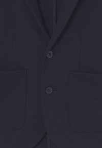 Námořnický modrý blazer s hladkou texturou, dvěma předními kapsami a třemi knoflíky. Čisté linie s přizpůsobeným střihem a jemnými detaily šití.