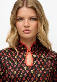 Schwarzes Kleidungsstück mit roten und gelben Blumenmustern, einem Ausschnitt in Schlüssellochform und rotem Besatz am Kragen. Glatte Textur.