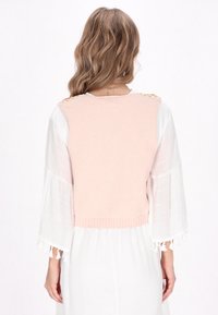 Rosa gestrickter kurz geschnittener Pullover mit dekorativen Akzenten an den Schultern; weiche Textur; kombiniert mit einem weißen, voluminösen Kleid mit Quasten-Details.
