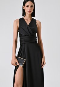 Zwart mouwloos jurkje met een wikkeldesign, diepe V-hals, zijsplit, gemaakt van gladde stof. Model houdt een kleine zwarte clutch met gouden accenten vast.