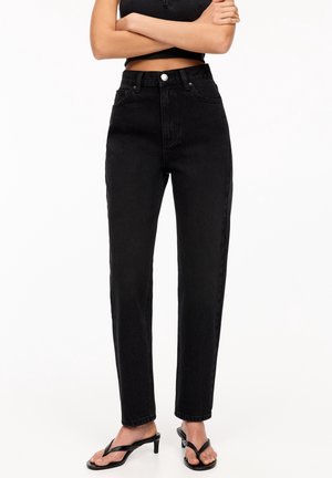 Kvinde iført sorte jeans med høj talje og lige ben, sort crop top og sorte hælede sandaler, med armene krydset, mod en ensfarvet hvid baggrund.
