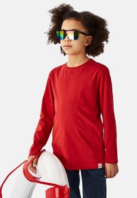 Rød langærmet skjorte lavet af bomuld med rund halsudskæring. Barnet holder en hvid redningskrans med et rødt reb og bærer reflekterende solbriller.
