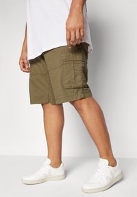 Olivgröna cargo shorts i bomull med sidofickor, i kombination med vita sneakers. Shortsen har en avslappnad passform och synliga sömmar.