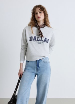 Giovane donna con capelli ricci che indossa una felpa grigia Dallas, jeans azzurri, sciarpa leopardata e tiene in mano una borsa nera.