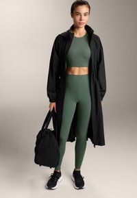 Čierny dlhý kabát cez zelený crop top a zodpovedajúce legíny. Čierne tenisky a čierny batoh s textúrovanou látkou. Neutrálne pozadie.