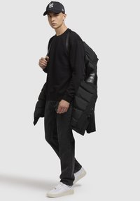 Sort pufferjakke båret over en sort sweatshirt og mørke jeans, suppleret med hvide sneakers og en sort kasket med et logo.