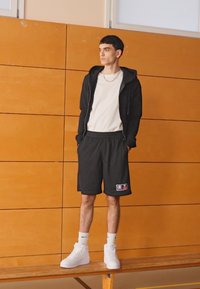 Pantalones cortos deportivos negros con bolsillos laterales y parche con logo, combinados con una camiseta blanca y una sudadera con cremallera negra. Zapatillas blancas de caña alta.