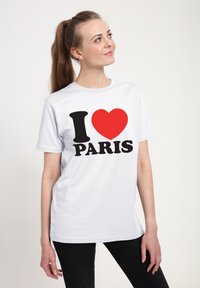 Bílá bavlněná trička s výrazným černým textem "I" a "PARIS" s červeným srdcem mezi nimi, nošena s černými kalhotami.
