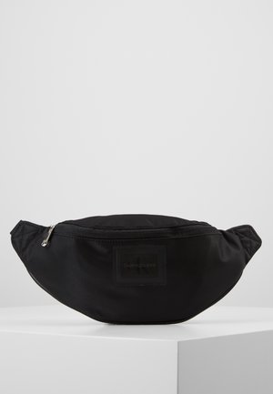 Musta nailonvyölaukku, jossa vetoketjusuljenta ja Calvin Klein Jeans -logokilpi edessä, esillä valkoisella alustalla.