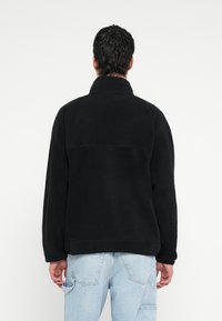 Czarna kurtka z polaru z wysokim kołnierzem, widocznymi przeszyciami i luźnym krojem, zestawiona z jasnoniebieskimi dżinsami.