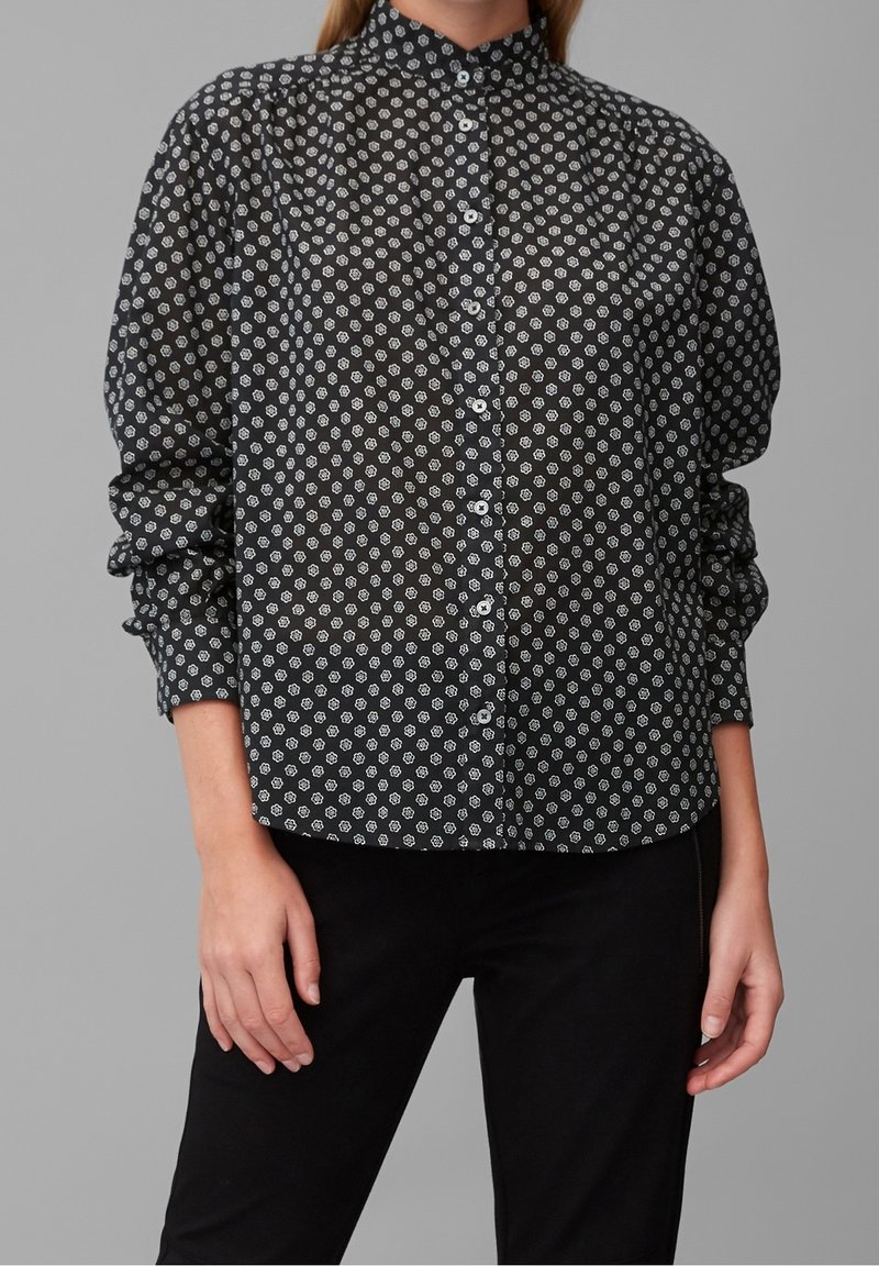 Femme portant une chemise noire à boutons avec de petits motifs floraux blancs et des manches longues, associée à un pantalon noir sur un fond gris.