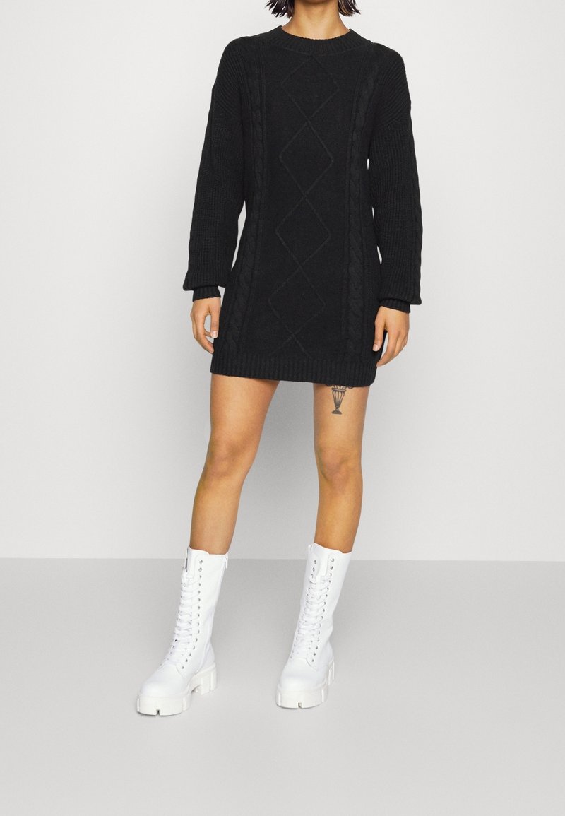 Personne portant une robe-pull noire en maille torsadée associée à des bottes épaisses blanches à lacets, debout les mains détendues le long du corps.