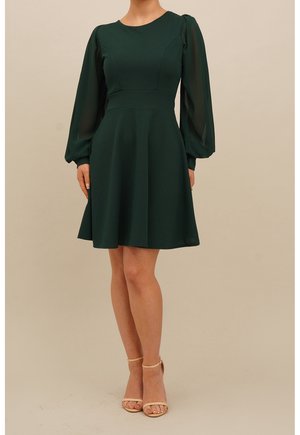 Mujer con vestido verde oscuro hasta la rodilla con mangas largas abullonadas transparentes y tacones beige con tiras en el tobillo, de pie contra un fondo sencillo.