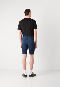 Shorts in denim blu scuro con vestibilità regolare, tasche posteriori, indossati con una t-shirt nera e scarpe da ginnastica nere, con calzini bianchi.